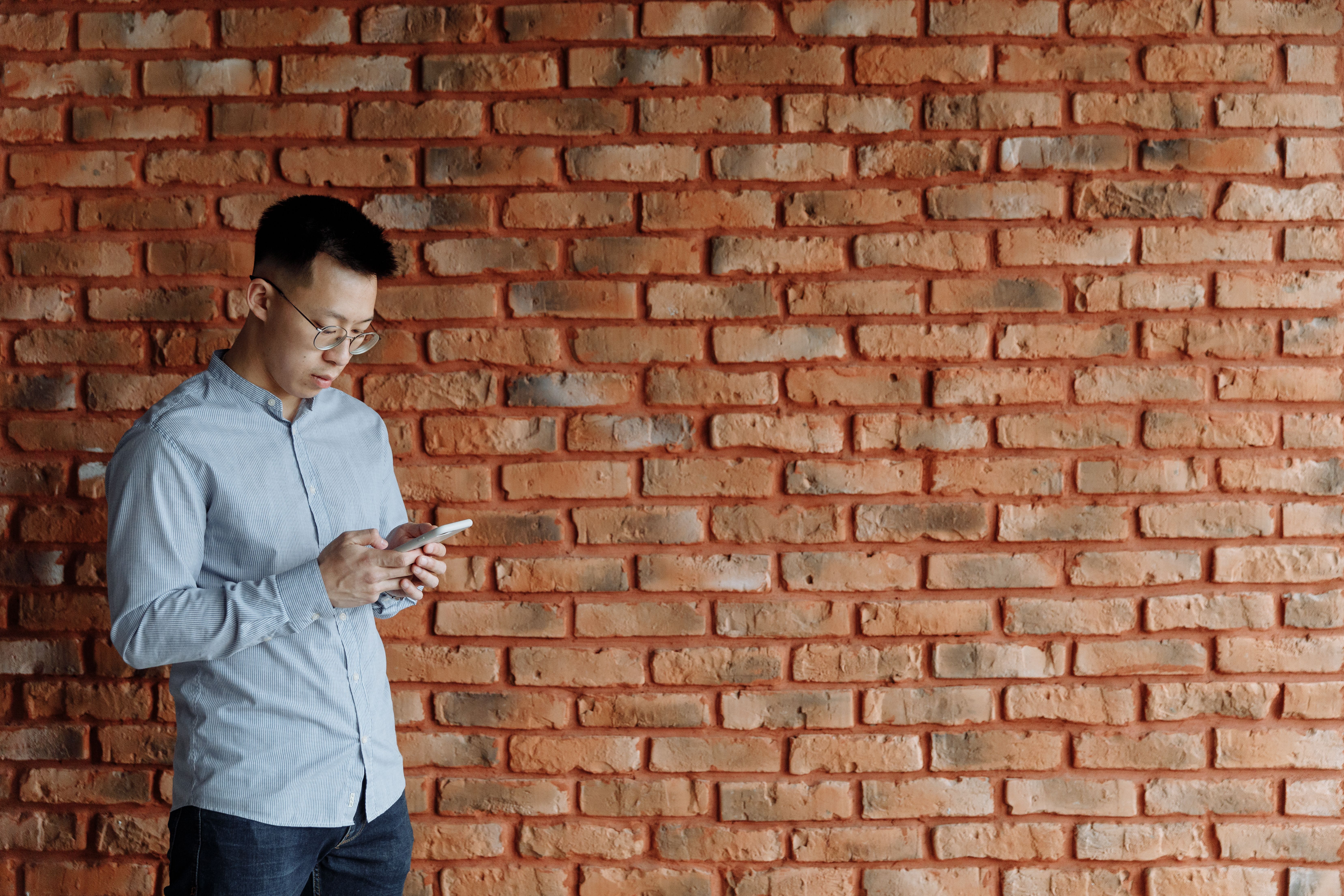 Asian man using a smartphone against a brick wall, representing social media use and misinformation spread